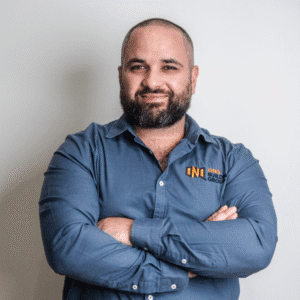 Brendan, Director of ONQ Building Group, smiling with arms crossed, wearing a blue shirt with company logo, representing leadership in quality home renovations and a family-first approach.